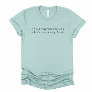 May include: A light blue t-shirt with the text "CAN'T THROW STONES while washing feet" printed in black.