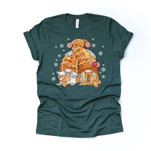 May include: A green t-shirt with a design of two gingerbread gnomes holding gingerbread men. The gnomes are wearing candy cane hats and have white beards. The design is surrounded by white snowflakes.
