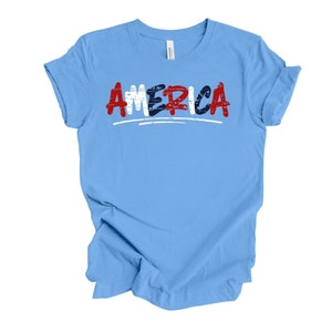 May include: Light blue t-shirt with the word "AMERICA" written in red, white, and blue with a distressed look.