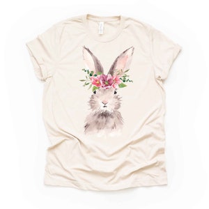 May include: A cream-colored t-shirt featuring a watercolor illustration of a bunny wearing a floral crown. The bunny has gray and brown fur, pink ears, and is adorned with pink flowers and green foliage. The shirt has short sleeves and a crew neck.