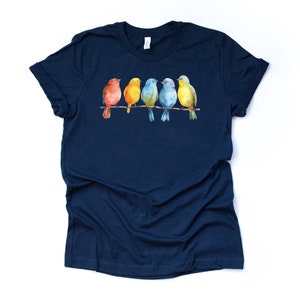 May include: A navy blue t-shirt with a watercolor design of five colorful birds perched on a branch. The birds are red, yellow, green, blue, and yellow.