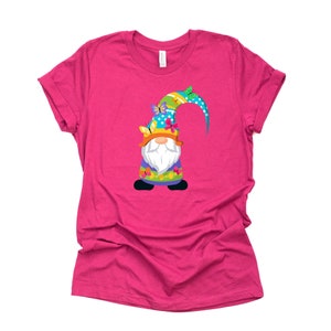 Könnte beinhalten: Ein pinkfarbenes T-Shirt mit einem bunten Zwerg, der einen blauen und grünen Hut mit Schmetterlingen trägt.