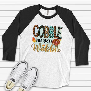 May include: A white and black baseball tee shirt with a Thanksgiving themed graphic that says "Gobble Till You Wobble" with a turkey illustration. The shirt has a black and white color block design.