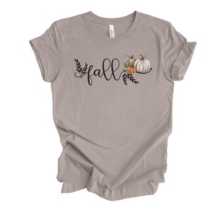 May include: A heather grey t-shirt with the word "fall" written in black script with a small cluster of pumpkins and leaves to the right of the word.