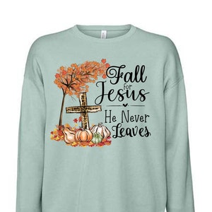 Puede incluir: Una sudadera de color azul claro con un dise&ntilde;o de oto&ntilde;o que incluye una cruz, calabazas y hojas. El texto dice "Fall for Jesus He Never Leaves".