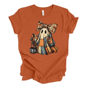 May include: Orange t-shirt featuring a cartoon ghost with a floral bow and patchwork design. The ghost is holding a jack-o'-lantern bucket. The design has orange, blue, and cream colors, perfect for Halloween.