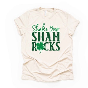 Puede incluir: Una camiseta beige con el texto "Shake Your Shamrocks" en letras verdes con un tr&eacute;bol de cuatro hojas verde.