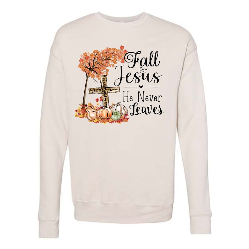 Puede incluir: Una sudadera blanca con un dise&ntilde;o de oto&ntilde;o que presenta una cruz, hojas y calabazas. El texto dice "Fall for Jesus He Never Leaves".