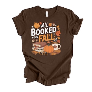 May include: Brown t-shirt with the text "All Booked for Fall" in white and orange. The design includes fall-themed illustrations such as leaves, a pumpkin, books, a coffee mug, and eyeglasses.