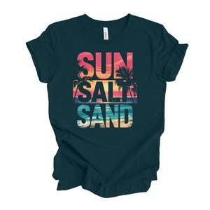 Op de afbeelding: Donker teal t-shirt met de woorden "SUN SALT SAND" in een kleurrijk, gelaagd ontwerp. De tekst bevat een zonsondergang en een palmboomafbeelding. Het shirt is gemaakt van een zacht materiaal.
