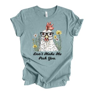 May include: A light blue t-shirt featuring a whimsical illustration of a chicken wearing glasses and surrounded by flowers. The text "Don't Make Me Peck You" is printed below the image. The shirt is made of a soft material.