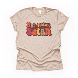 May include: A beige t-shirt with the text "Not today, Satan" in colorful retro-style lettering.
