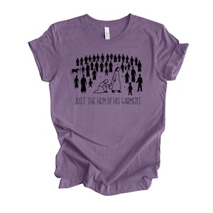 May include: Purple t-shirt with a black graphic of a crowd of people, a kneeling figure, and the text "JUST THE HEM OF HIS GARMENT." The shirt is made of a soft material and has a crew neck.