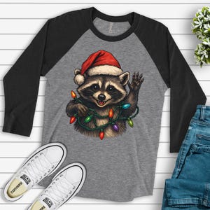 May include: A gray raglan-style shirt with black sleeves. The shirt features a raccoon wearing a Santa hat and a string of Christmas lights. White Converse sneakers and blue jeans are also visible.