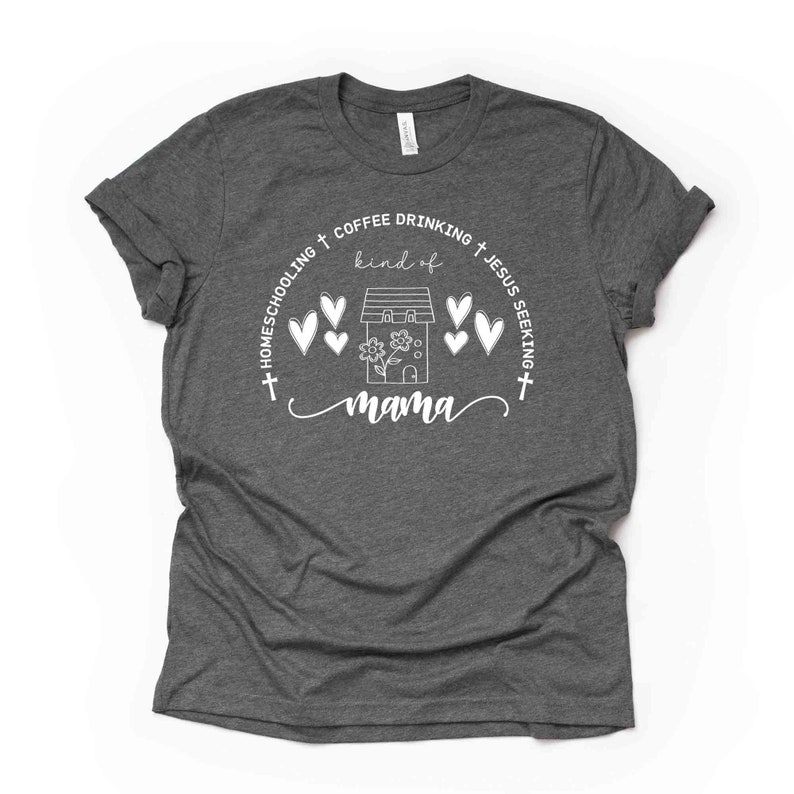 Puede incluir: Una camiseta gris oscuro con un dise&ntilde;o gr&aacute;fico blanco. El dise&ntilde;o presenta una casa con corazones, flores y el texto "Homeschooling + Coffee Drinking + Jesus Seeking + kind of mama".