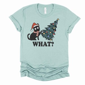 May include: A light blue t-shirt with a black cat wearing a Santa hat and scarf, knocking over a green Christmas tree with colorful ornaments. The text "WHAT?" is printed below the image.