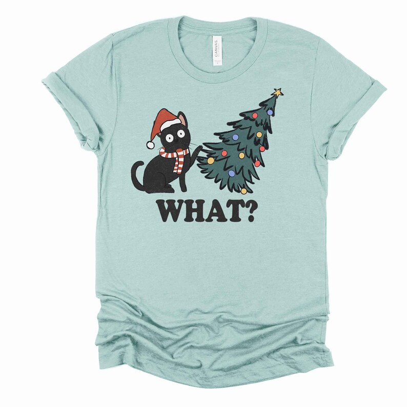 May include: A light blue t-shirt with a black cat wearing a Santa hat and scarf, knocking over a green Christmas tree with colorful ornaments. The text "WHAT?" is printed below the image.