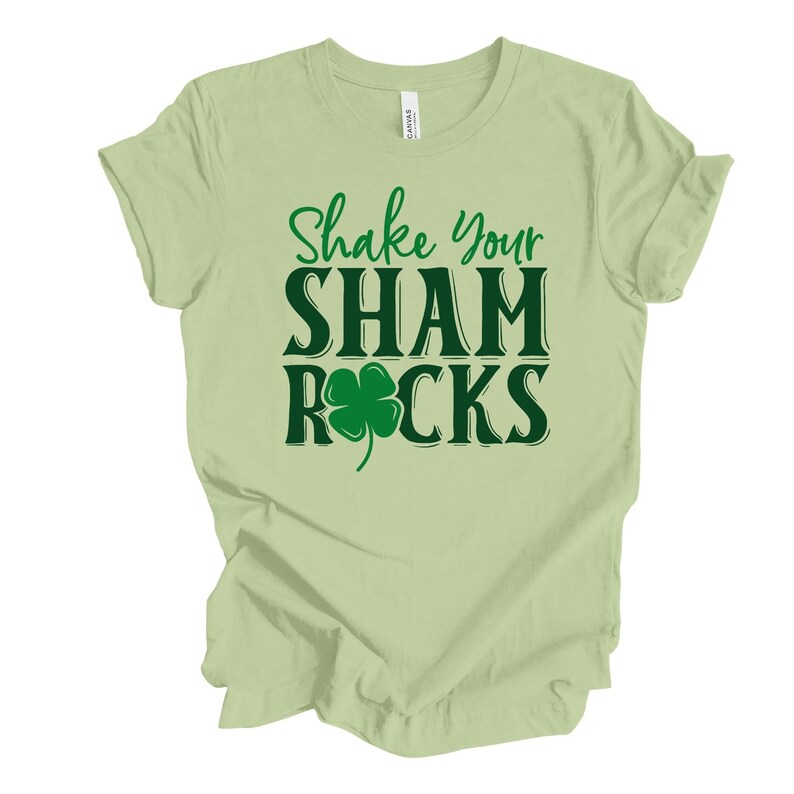 Puede incluir: Una camiseta verde claro con el texto "Shake Your Shamrocks" en verde oscuro y un gr&aacute;fico de tr&eacute;bol verde.