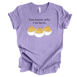 May include: A light purple t-shirt with the text "You know why I'm here." and a graphic of three deviled eggs. The deviled eggs have a yellow-orange filling and are in white eggshell halves.