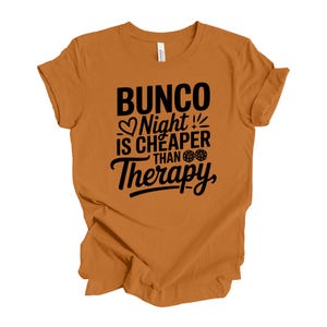 K&ouml;nnte beinhalten: Ein orangefarbenes T-Shirt mit dem schwarzen Text "Bunco Night is Cheaper Than Therapy". Das Design umfasst ein Herz, W&uuml;rfel und Blitze. Das Shirt besteht aus einem weichen Material.
