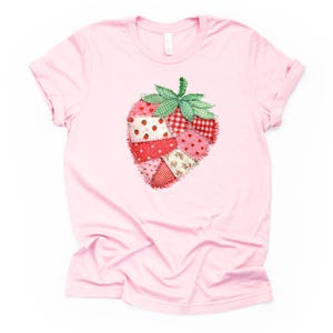 May include: A light pink t-shirt featuring a patchwork strawberry design. The strawberry is composed of various fabric patterns in shades of red, pink, and white, with green leaves. The shirt has short sleeves and a crew neck.