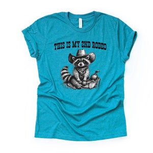 Pode incluir: Uma camiseta azul turquesa com uma ilustra&ccedil;&atilde;o em preto e branco de um guaxinim usando um chap&eacute;u de cowboy e montando uma sela. O texto "This is my 2nd Rodeo" est&aacute; impresso na camiseta.