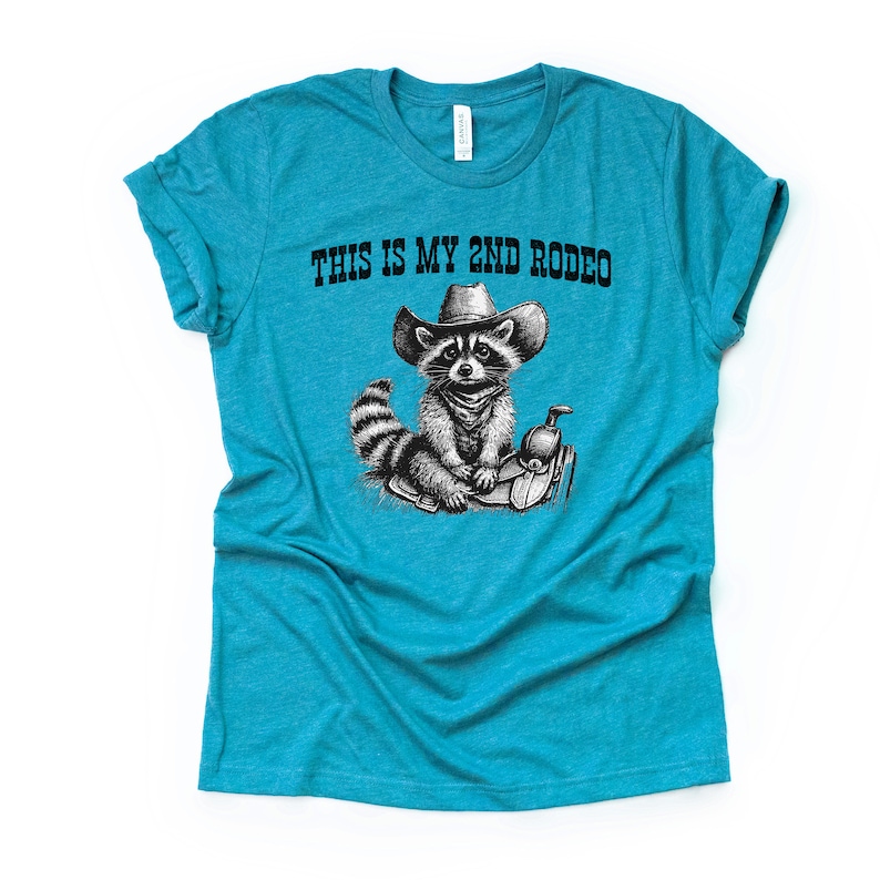 Pode incluir: Uma camiseta azul turquesa com uma ilustra&ccedil;&atilde;o em preto e branco de um guaxinim usando um chap&eacute;u de cowboy e montando uma sela. O texto "This is my 2nd Rodeo" est&aacute; impresso na camiseta.