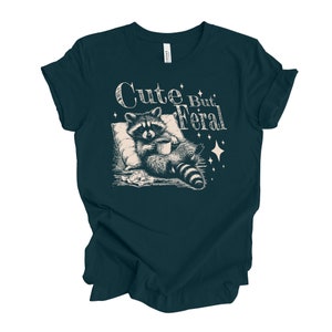 May include: A dark teal t-shirt with a white graphic of a raccoon holding a cup of coffee and the text "Cute But Feral".