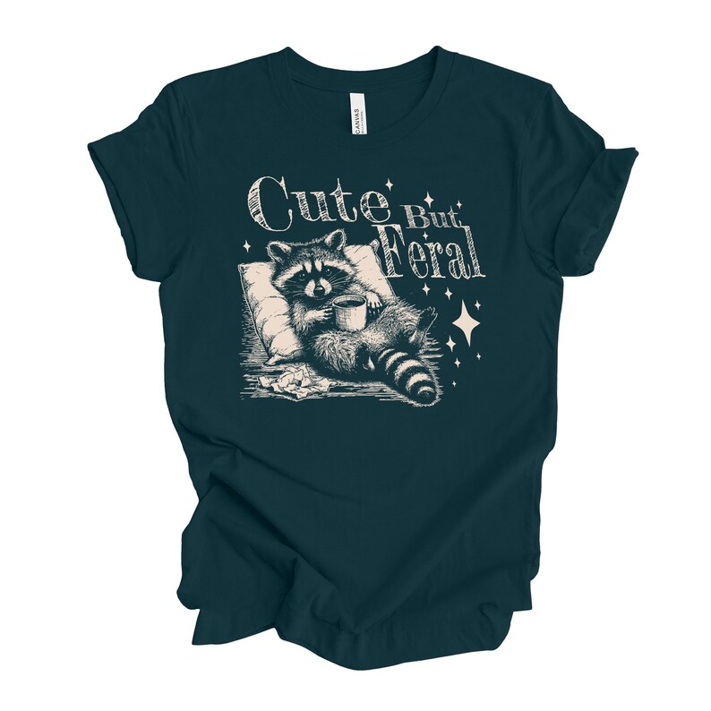 May include: A dark teal t-shirt with a white graphic of a raccoon holding a cup of coffee and the text "Cute But Feral".