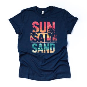 Op de afbeelding: Marineblauw T-shirt met de woorden "SUN SALT SAND" in een kleurrijk ontwerp. De tekst bevat een zonsondergang en palmbomen, met een strandsc&egrave;ne aan de onderkant. Een casual t-shirt voor strandliefhebbers.