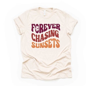 May include: A beige t-shirt with the text "FOREVER CHASING SUNSETS" in a retro font with a flower graphic. The text is in shades of burgundy, orange, and yellow.