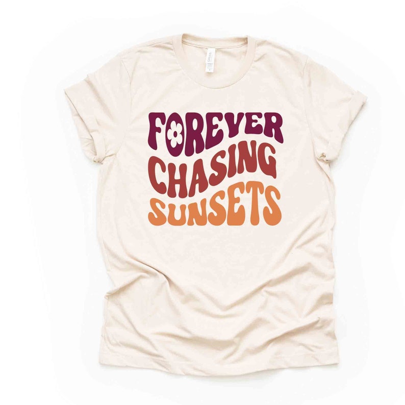 May include: A beige t-shirt with the text "FOREVER CHASING SUNSETS" in a retro font with a flower graphic. The text is in shades of burgundy, orange, and yellow.