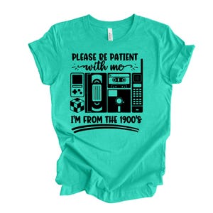 May include: A turquoise t-shirt with a black graphic design featuring retro electronics like a handheld game console, VHS tape, cassette tape, and a brick phone. The text reads "PLEASE BE PATIENT with me I'M FROM THE 1900's."
