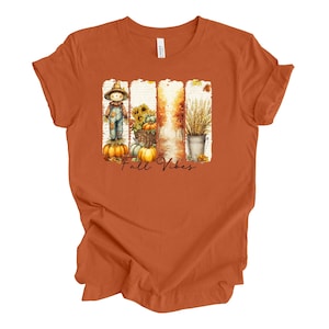 May include: Orange t-shirt with a fall-themed graphic. The graphic features a scarecrow, pumpkins, sunflowers, and wheat, with the words "Fall Vibes" in a script font. The shirt is made of a soft material.
