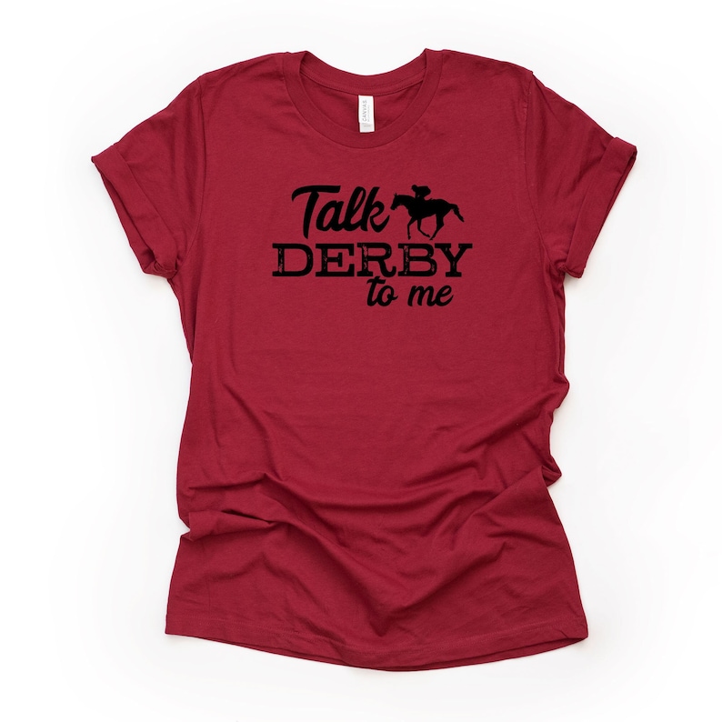 Peut inclure: T-shirt rouge avec un graphisme noir qui dit "Talk Derby to me" et une silhouette de jockey &agrave; cheval.