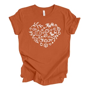 May include: A burnt orange short-sleeve t-shirt featuring a white graphic print of a heart shape composed of various autumn-themed line art illustrations, including pumpkins, acorns, leaves, flowers, and small branches on the chest area.