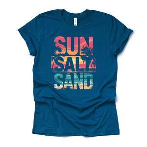 Op de afbeelding: Een tealkleurig t-shirt met de woorden "SUN SALT SAND" in een kleurrijk strandthema. De tekst is gelaagd met een zonsondergang en een palmboomafbeelding, die een kustsfeer oproept. Het shirt is gemaakt van een zacht materiaal.