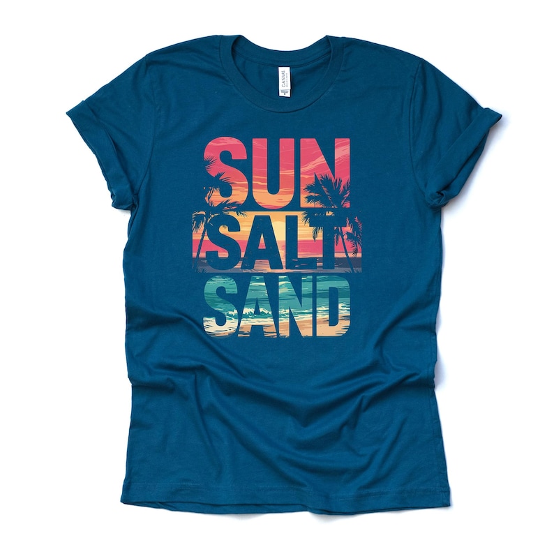 Op de afbeelding: Een tealkleurig t-shirt met de woorden "SUN SALT SAND" in een kleurrijk strandthema. De tekst is gelaagd met een zonsondergang en een palmboomafbeelding, die een kustsfeer oproept. Het shirt is gemaakt van een zacht materiaal.