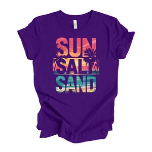 Op de afbeelding: Een paars t-shirt met de woorden "SUN SALT SAND" in een kleurrijk strandthema. De tekst is over een zonsondergang en palmbomen geplaatst, wat een vakantiegevoel oproept.