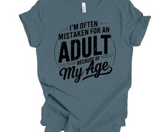Puede incluir: Una camiseta azul con el texto negro "I'M OFTEN MISTAKEN FOR AN ADULT BECAUSE OF My Age". El texto está dispuesto en un diseño circular. La camiseta es de material suave y tiene mangas cortas.