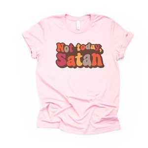 May include: Pink t-shirt with the text "Not today, Satan" in retro-style lettering with orange, red, brown, and beige colors.