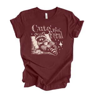 May include: A maroon t-shirt with a white graphic of a raccoon holding a cup of coffee and the text "Cute But Feral".