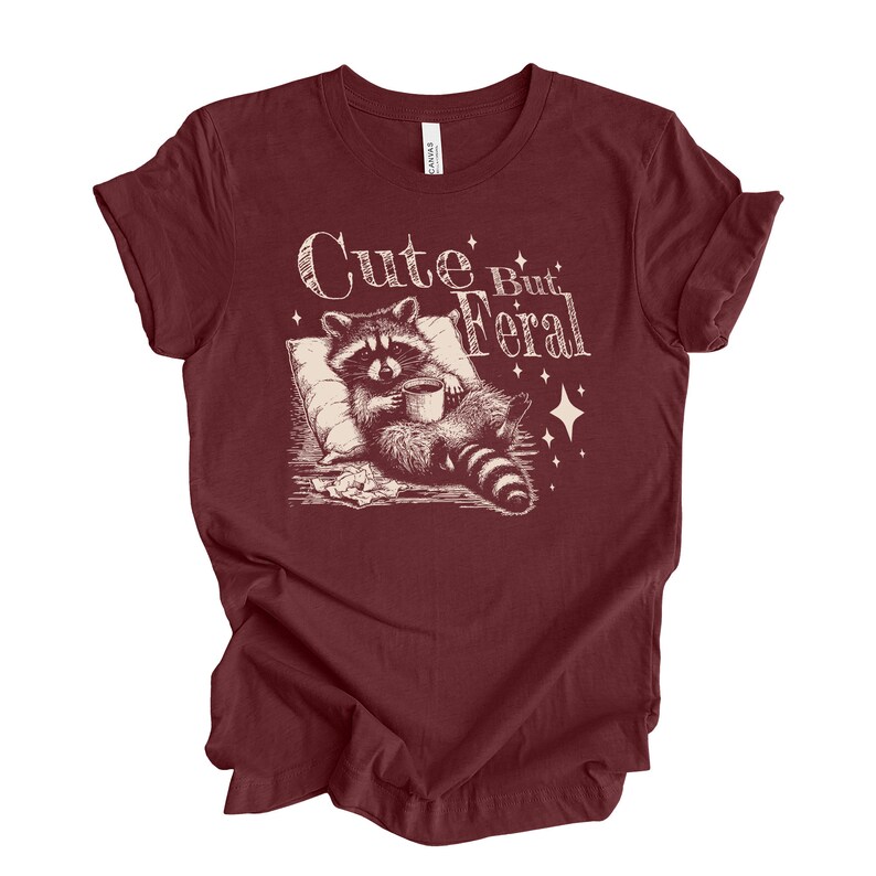 May include: A maroon t-shirt with a white graphic of a raccoon holding a cup of coffee and the text "Cute But Feral".