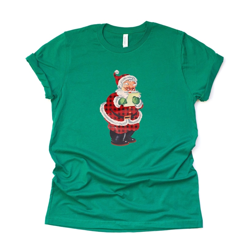 May include: A green t-shirt with a vintage illustration of Santa Claus wearing a red and black plaid suit and holding a scroll.
