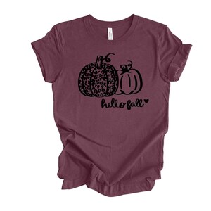 May include: Burgundy t-shirt featuring a black graphic of two pumpkins, one with a leopard print design. The words "hello fall" are written in a script font below the pumpkins, with a small heart symbol. The shirt is made of a soft material.