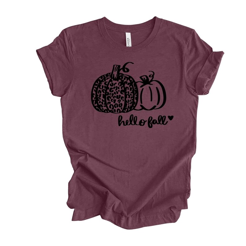 May include: Burgundy t-shirt featuring a black graphic of two pumpkins, one with a leopard print design. The words "hello fall" are written in a script font below the pumpkins, with a small heart symbol. The shirt is made of a soft material.