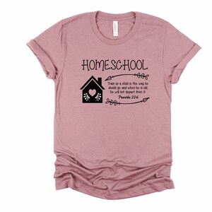 Puede incluir: Una camiseta rosa con el texto "HOMESCHOOL" y una ilustraci&oacute;n negra de una casa con un coraz&oacute;n y flores. El texto "Train up a child in the way he should go and when he is old, he will not depart from it. Proverbs 22:6" tambi&eacute;n est&aacute; impreso en la camiseta.