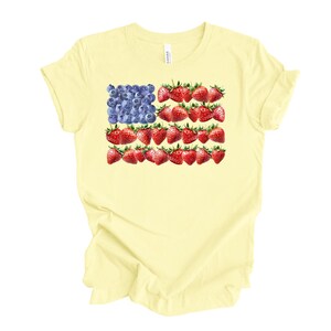 May include: A light yellow t-shirt featuring a patriotic design. The design depicts the American flag, with blueberries replacing the stars and strawberries replacing the stripes. The shirt is a casual wear item.
