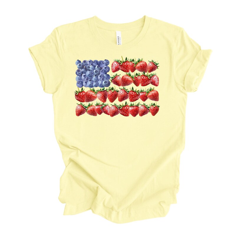 May include: A light yellow t-shirt featuring a patriotic design. The design depicts the American flag, with blueberries replacing the stars and strawberries replacing the stripes. The shirt is a casual wear item.