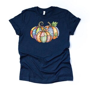 Pu&ograve; includere: Una maglietta blu navy con una grafica di tre zucche. Le zucche hanno un design patchwork con vari motivi e colori, tra cui pois, cuori e strisce. Le zucche sono disposte in fila, con foglie verdi e steli ricci.